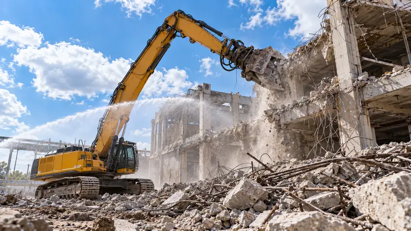 A high-reach excavator demolishing a concrete building, with a water spray keeping the dust down